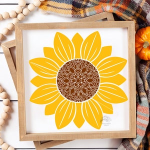 K&ouml;nnte beinhalten: Ein quadratischer Holzrahmen mit einem wei&szlig;en Schild, das eine gro&szlig;e gelbe Sonnenblumenillustration zeigt. Die Sonnenblume hat eine detaillierte braune Mitte im Mandala-Stil mit komplexen wei&szlig;en Linienmustern. Die Bl&uuml;tenbl&auml;tter sind einfarbig gelb.