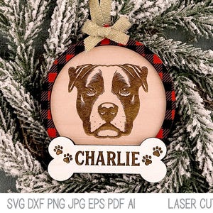 May include: A wooden Christmas ornament featuring a dog's head with the name "CHARLIE" on a bone-shaped tag. The ornament is decorated with a red and black buffalo plaid pattern.