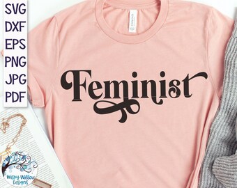 May include: A light peach-colored t-shirt featuring the word Feminist printed in a bold, dark brown, stylized serif font across the chest. The shirt has a crew neckline and short sleeves, with a visible brand label inside the collar area.