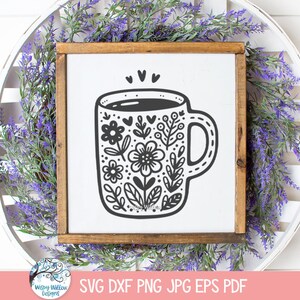 Op de afbeelding: Een zwart-witte illustratie van een koffiemok met een bloemenontwerp. De mok is gevuld met koffie en heeft een hart erboven.