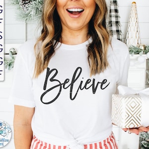 Könnte beinhalten: Ein weißes T-Shirt mit dem Wort "Believe" in schwarzer Kursivschrift. Das Shirt wird von einer Person mit langen braunen Haaren getragen und hält eine verpackte Geschenkbox mit einem gold-weißen Muster.