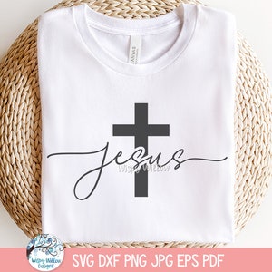May include: A white short-sleeved t-shirt featuring a dark gray graphic design of a vertical cross intersected by the word Jesus in a cursive script font. The shirt has a crew neckline and a visible brand label inside the collar area.