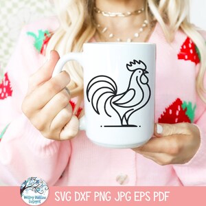 May include: A white ceramic mug with a black line drawing of a rooster on it. The mug is being held by a person wearing a pink sweater with a strawberry pattern.
