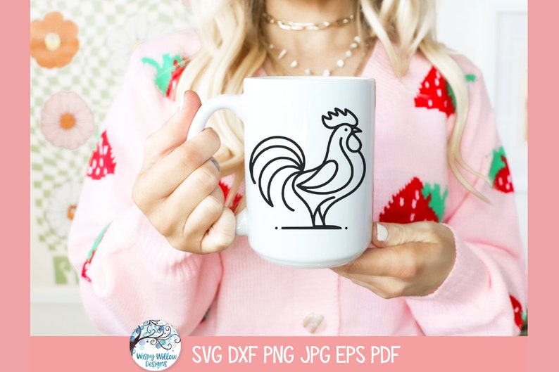 May include: A white ceramic mug with a black line drawing of a rooster on it. The mug is being held by a person wearing a pink sweater with a strawberry pattern.