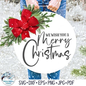 May include: A round white sign featuring the black text "WE WISH YOU A Merry Christmas" in a mix of block and script fonts. The sign is decorated with a large red burlap bow, green leaves, and pine branches, hanging from a rope handle.