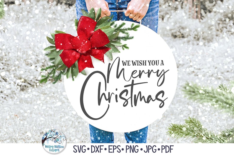 May include: A round white sign featuring the black text "WE WISH YOU A Merry Christmas" in a mix of block and script fonts. The sign is decorated with a large red burlap bow, green leaves, and pine branches, hanging from a rope handle.