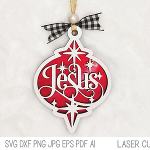 May include: A teardrop-shaped Christmas ornament featuring a white cutout frame with the word Jesus in script, layered over a red background with star accents. It is topped with a black and white buffalo plaid bow and a gold hanging cord.