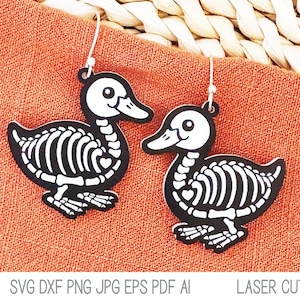 May include: A pair of skeleton duck earrings with black and white details. The earrings are hanging from a woven basket on a peach-colored fabric background.