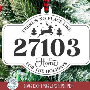 May include: A white, rectangular, rounded-edge Christmas ornament featuring the black text 'THERE'S NO PLACE LIKE 27103 HOME FOR THE HOLIDAYS' with a central graphic of a leaping reindeer between two pine trees, suspended by a red and black plaid ribbon.