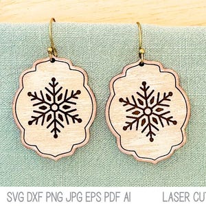 May include: A pair of wooden snowflake earrings with a laser cut design. The earrings are a light brown color and have a gold-colored metal hook.
