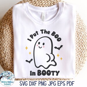 以下が含まれることがあります： ピンクの頬をしたゴーストの黒いグラフィックが描かれた白いTシャツ。黒い文字で「I Put The BOO In BOOTY」と書かれています。デザインには、小さな黄色い四角い星と、ゴーストの図の両側に2匹の黒いコウモリが含まれています。