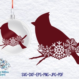 May include: A clear glass Christmas ornament featuring a dark red cardinal silhouette perched on a branch with white snowflake and flower accents, accompanied by a matching dark red cardinal graphic with white snowflakes and the text Wispy Willow Designs.