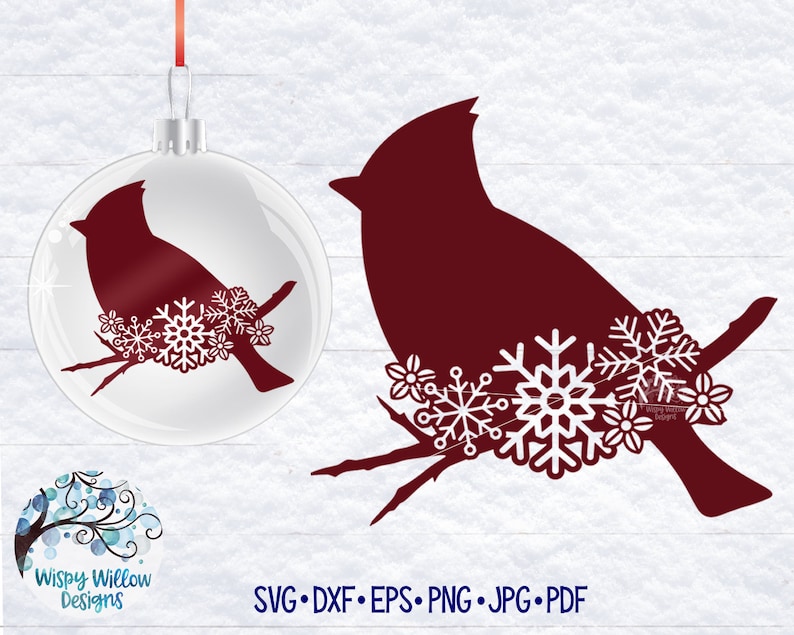 May include: A clear glass Christmas ornament featuring a dark red cardinal silhouette perched on a branch with white snowflake and flower accents, accompanied by a matching dark red cardinal graphic with white snowflakes and the text Wispy Willow Designs.