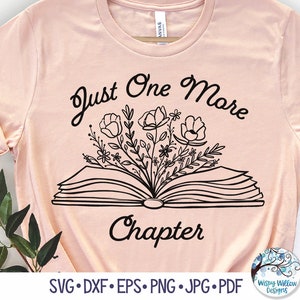 May include: A light peach-colored t-shirt featuring a black line-art graphic of an open book with flowers emerging from the pages. The text "Just One More Chapter" is printed in black above and below the book illustration on the front of the shirt.