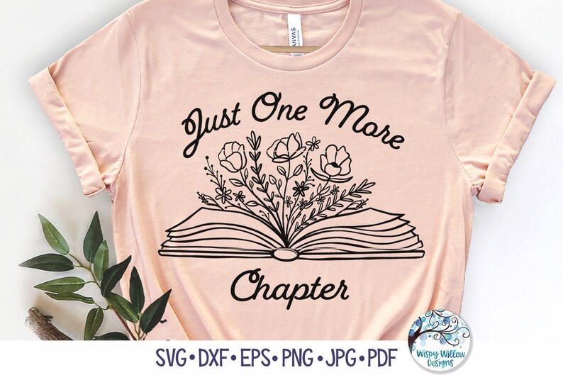 May include: A light peach-colored t-shirt featuring a black line-art graphic of an open book with flowers emerging from the pages. The text "Just One More Chapter" is printed in black above and below the book illustration on the front of the shirt.