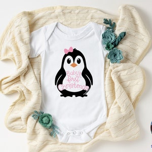 May include: White baby onesie with a black and white penguin graphic wearing a pink bow. The text "Baby's first Christmas" is in pink script. The onesie is on a cream-colored blanket with teal floral accents.