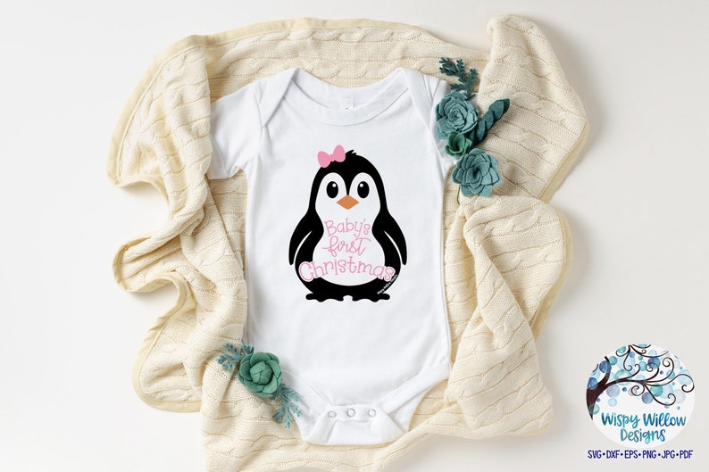 May include: White baby onesie with a black and white penguin graphic wearing a pink bow. The text "Baby's first Christmas" is in pink script. The onesie is on a cream-colored blanket with teal floral accents.