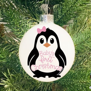May include: White Christmas ornament featuring a black and white penguin with a pink bow and the text "Baby's first Christmas." The ornament is hanging on a Christmas tree with green needles and brown pinecones.