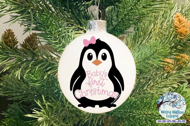 May include: White Christmas ornament featuring a black and white penguin with a pink bow and the text "Baby's first Christmas." The ornament is hanging on a Christmas tree with green needles and brown pinecones.