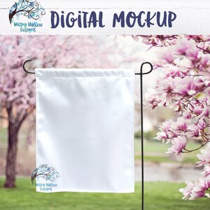 May include: White blank garden flag mockup with a floral background. The flag is hanging from a black metal pole. The flag is 12 inches by 18 inches. The background is a blurred image of a pink magnolia tree.