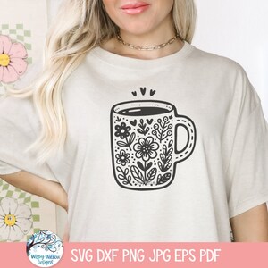 Op de afbeelding: Een wit T-shirt met een zwart-witte illustratie van een koffiemok met bloemenmotieven. De mok heeft een zwarte omtrek en is gevuld met een zwarte vloeistof. De bloemenmotieven zijn ingewikkeld en bevatten bladeren, bloemen en harten.