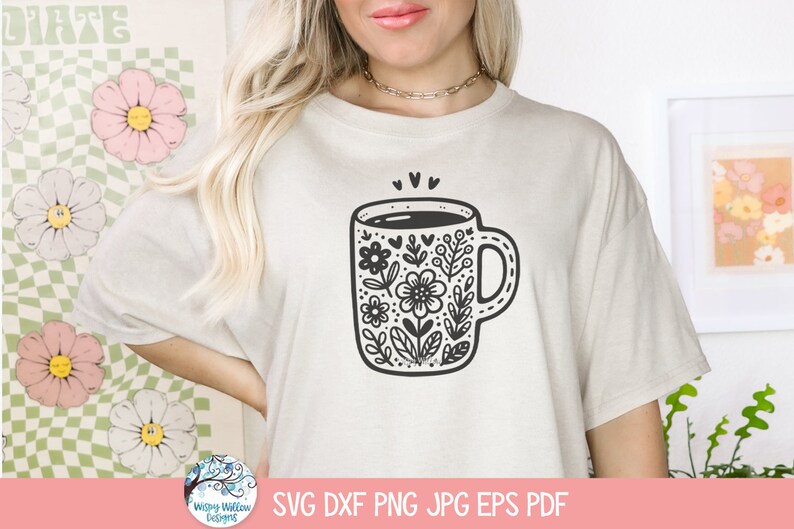 Op de afbeelding: Een wit T-shirt met een zwart-witte illustratie van een koffiemok met bloemenmotieven. De mok heeft een zwarte omtrek en is gevuld met een zwarte vloeistof. De bloemenmotieven zijn ingewikkeld en bevatten bladeren, bloemen en harten.