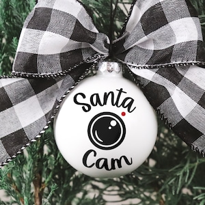 May include: A white spherical Christmas ornament featuring the black text Santa Cam and a black camera lens graphic with a small red dot. The ornament is topped with a large black and white buffalo plaid fabric bow tied with a black string.
