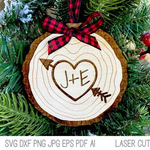 Op de afbeelding: Een houten kerstversiering in de vorm van een boomstam met een hart- en pijlontwerp. Het hart heeft de initialen "J+E" erin. De versiering is versierd met een rood-zwart geruit lint.