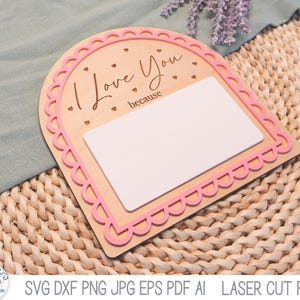 May include: An arched wooden sign with a pink scalloped border and the text "I Love You because" engraved in black. The sign features a large, blank white rectangular space for writing and small engraved heart accents on the light wood surface.