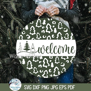 May include: A round dark green wooden sign featuring a white pattern of stylized pine trees and abstract shapes. A horizontal white band across the center displays the word welcome in black script, accompanied by three small tree icons.