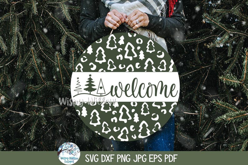 May include: A round dark green wooden sign featuring a white pattern of stylized pine trees and abstract shapes. A horizontal white band across the center displays the word welcome in black script, accompanied by three small tree icons.