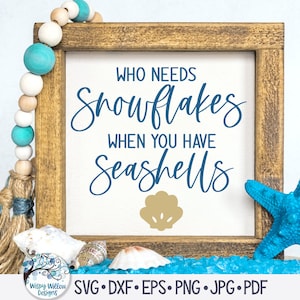 May include: A framed sign with the text "WHO NEEDS Snowflakes WHEN YOU HAVE Seashells" in blue script font. A gold seashell graphic is below the text. The sign is on a wooden surface with blue glass beads, a blue starfish, and seashells.