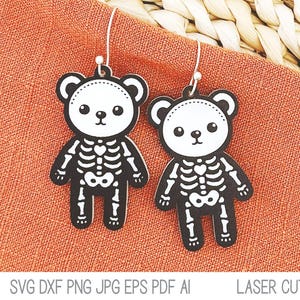 May include: A pair of dangle earrings featuring flat, black and white bear-shaped pendants with a printed skeleton design. Each bear has a white face with black eyes and a nose, and a black body with a white ribcage and pelvic bone graphic.