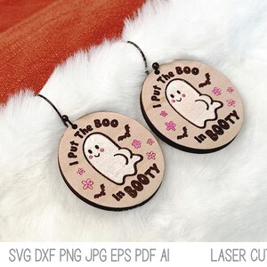 May include: A pair of round, light-colored wooden earrings featuring a laser-engraved cartoon ghost, small bats, and pink flowers. The text I Put The Boo In BOOTY is printed in black around the ghost. Each earring has a black metal hook.