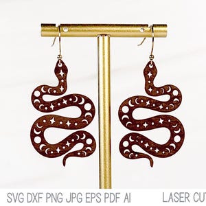 May include: A pair of laser cut wooden earrings shaped like a snake with a moon and star pattern. The earrings are brown and have silver hooks.