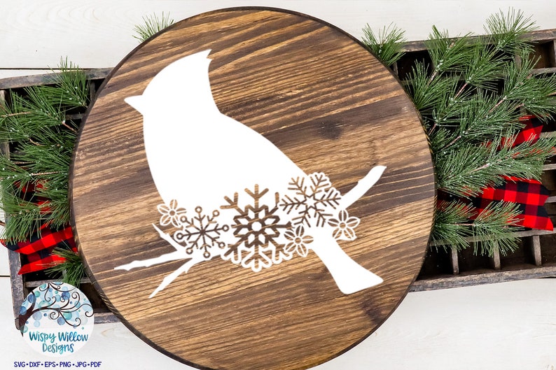 May include: A circular dark brown stained wooden sign featuring a white silhouette of a bird perched on a branch. The bird is decorated with intricate white snowflake and flower cutouts across its lower body. The design is flat and graphic.