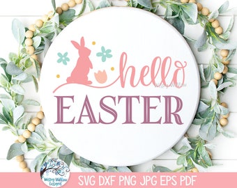 Pode incluir: Uma placa circular branca com a silhueta rosa de um coelho, flores azul-claras e um ovo cor de pêssego. O texto diz hello em letra cursiva rosa e EASTER em letras maiúsculas roxas grandes. O design é rodeado por uma coroa de folhas verdes.