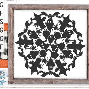 Op de afbeelding: Zwart-witte silhouet van een Halloween mandala-ontwerp met geesten, vleermuizen, spinnen en heksenhoeden. Het ontwerp is ingelijst in een rustiek houten frame.