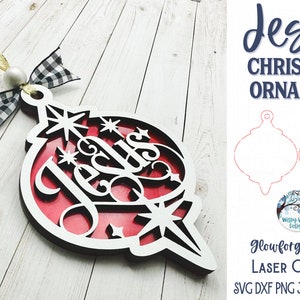 May include: A layered Christmas ornament featuring a black cutout frame with the word Jesus and star shapes over a solid red background. It includes a white bead, a black and white buffalo check ribbon, and a gold ribbon loop for hanging.