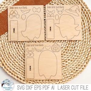 May include: Three wooden laser cutouts with the text "Build your own ghost". Each cutout features a ghost shape and various accessories like hats, bows, and pumpkin faces. The cutouts are on a woven mat with a brown fabric accent.