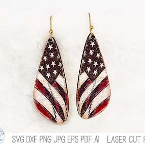 May include: A pair of teardrop-shaped earrings featuring a distressed American flag design with white stars on a dark background and red and white stripes. The earrings are suspended from gold-toned metal hooks and have a textured, weathered finish.