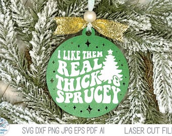 May include: A round green Christmas ornament featuring the white text I LIKE THEM REAL THICK & SPRUCEY and a white pine tree graphic. The ornament is topped with a small white bead and a gold glitter ribbon bow for hanging purposes.