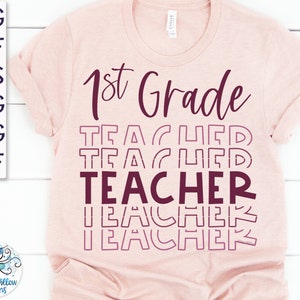 May include: A pink t-shirt with the text "1st Grade" and "TEACHER" in a layered, stacked design. The text is in a burgundy color.