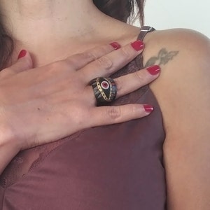 May include: A wide, dark brown ring with a black and red eye design. The ring has gold accents around the eye and is worn on a person's finger.
