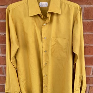 May include: Mustard yellow button-down shirt with a pointed collar, long sleeves, and a chest pocket. The shirt has a classic design and is hanging on a clear hanger. The brand name is visible on the collar.