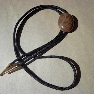 May include: A bolo tie featuring a brown, oval-shaped stone with a striped pattern. The stone is connected to a black braided cord with a gold-colored metal clasp.