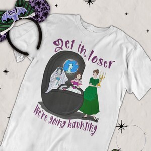 May include: White t-shirt with a cartoon illustration of three women in a coffin, with the text "Get in loser, we're going haunting." Also shown are a pair of purple and green bat-themed mouse ears and a silver bracelet with a charm.