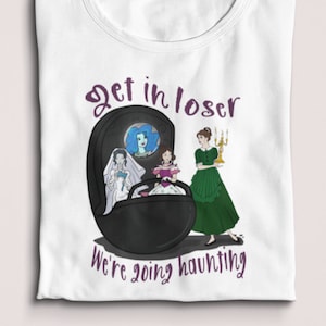 May include: White t-shirt with a graphic featuring a black oval with three figures and the text "Get in loser, we're going haunting" in purple. The figures include a bride, a woman in a green dress, and a woman in a blue dress.