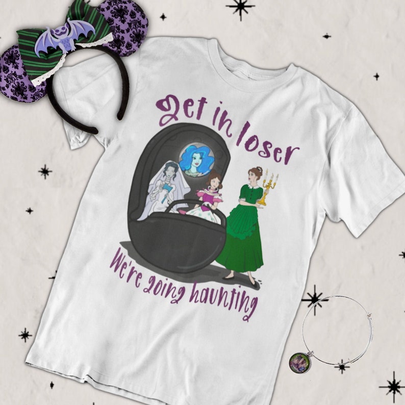 May include: White t-shirt with a cartoon illustration of three women in a coffin, with the text "Get in loser, we're going haunting." Also shown are a pair of purple and green bat-themed mouse ears and a silver bracelet with a charm.