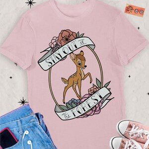 Könnte beinhalten: Hellrosa T-Shirt mit einem Cartoon-Hirsch-Design in einem goldenen Oval, mit dem Text "Stay Out of the Forest". Das Design umfasst Blumen und Banner. Rosa Turnschuhe und blaue Jeans sind sichtbar.